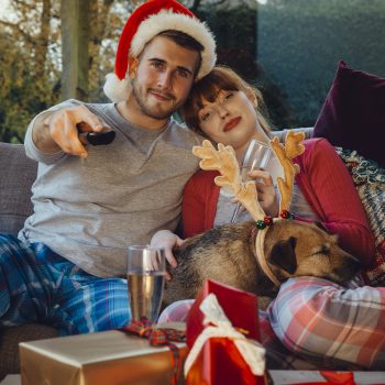 young couple wearing pajamas sitting on the couch with pet dog wearing reindeer antlers. man holding a remote wearing a santa hat