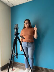 Kali Reaves films a vertical video using a tripod and an iPhone against a teal wall.