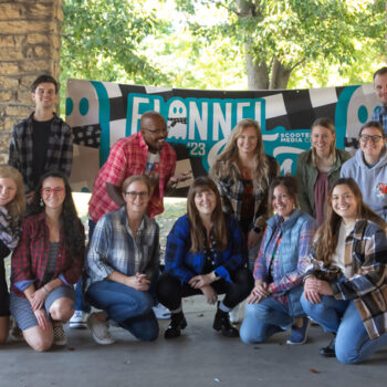 The Scooter Media team posing for a photo in front of a teal and black Flannel Fest banner.