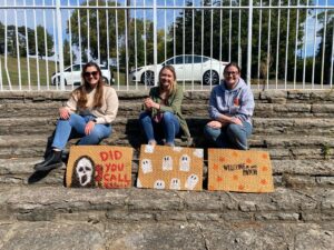 Three women posing on a set of outdoor stairs next to painted door rugs with Halloween designs at our annual Flannel Fest event.