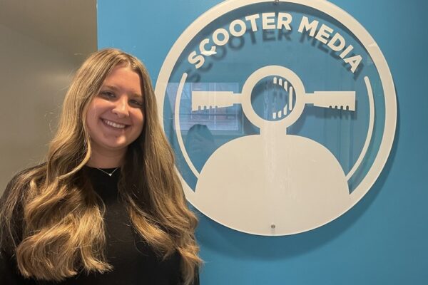Abbie Proctor, Assistant Account Executive, stands in front of the Scooter Media logo on a blue wall.