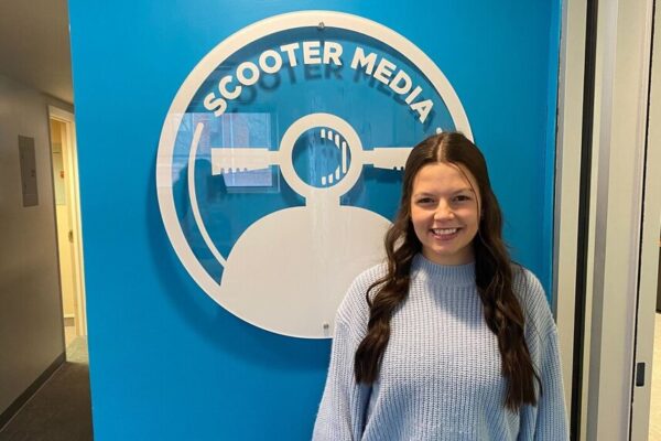 Nevaeh Votel, Assistant Account Executive, stands in front of the Scooter Media logo on a blue wall.